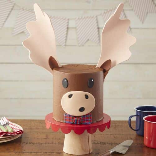 A round, brown cake as the face of a moose, topped with brown ears and big, beige antlers. Face is on the front of the cake, with a red and blue plaid bow tie.