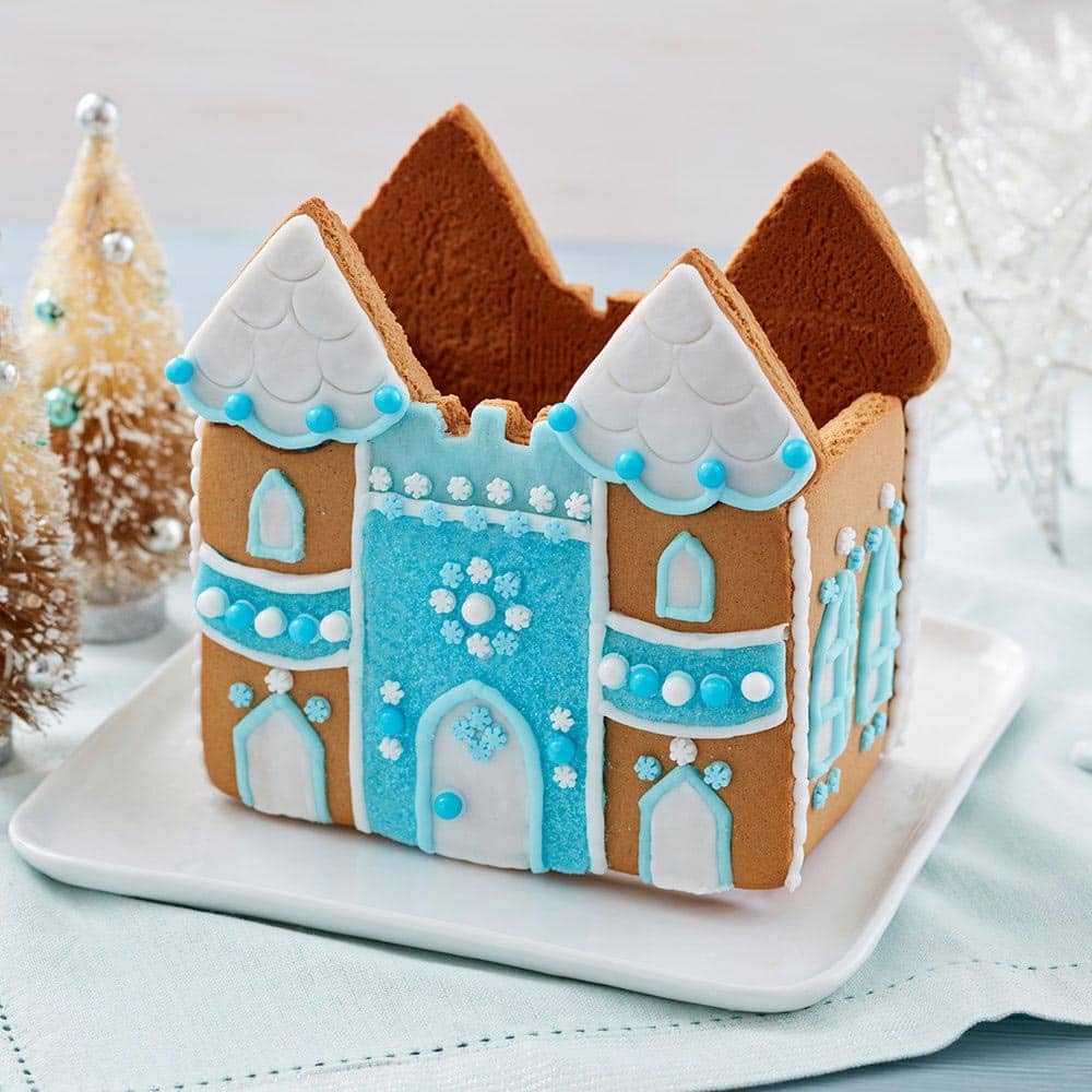 Gingerbread castle with a blue front and white roof. White doors and windows, outlined with blue icing. White and blue round sprinkles decorate the front and sides of the castle.