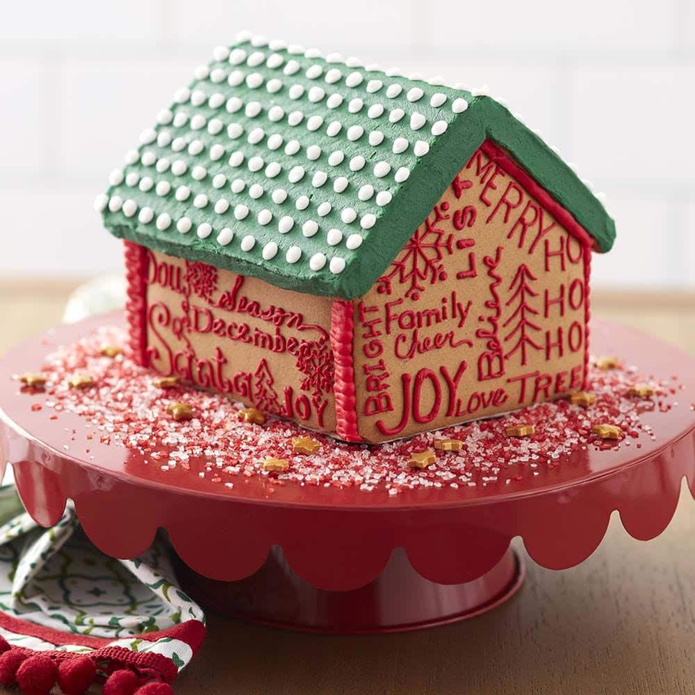 Gingerbread house with a green roof with white candy dots and on the walls of the house, Christmas words written in red. Words such as Joy, Family Cheer, Ho Ho Ho