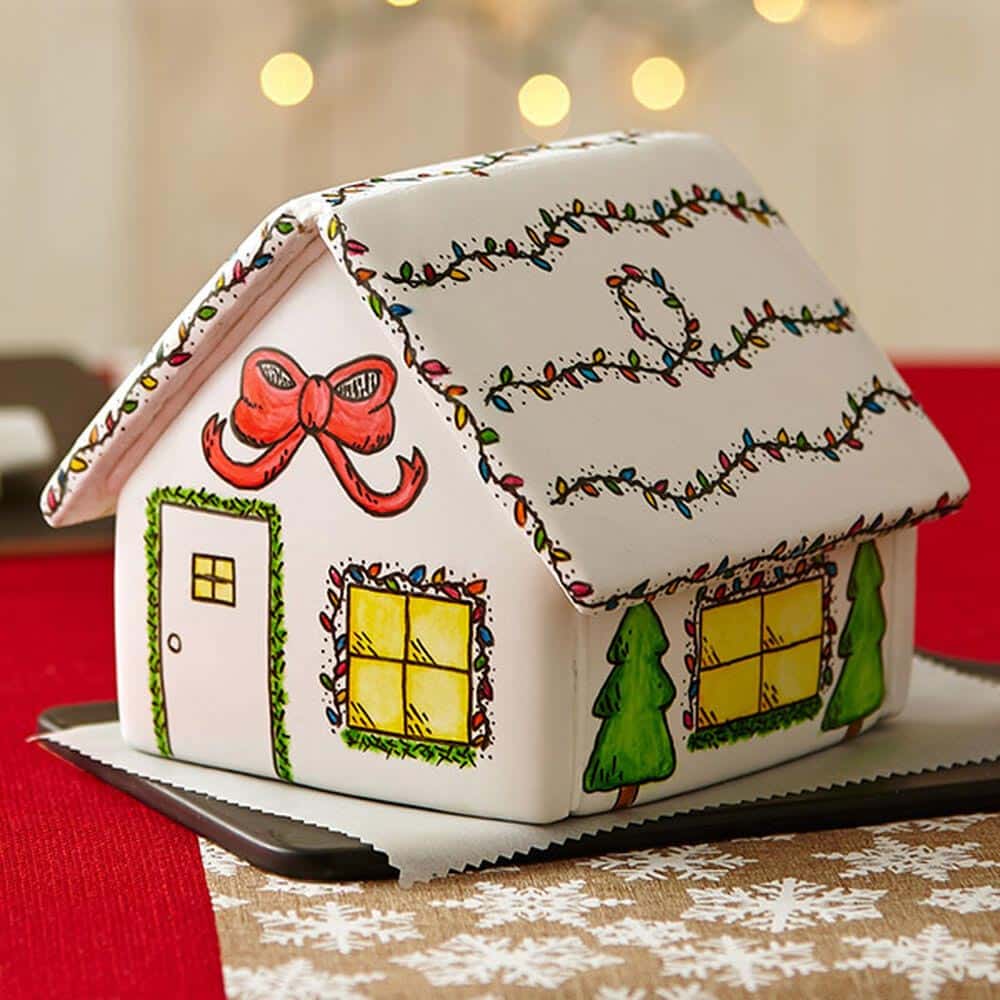 White gingerbread house with drawings for decorations. A big red bow sits over the front door, christmas lights line the roof, and two evergreens are on the side on either side of a window.