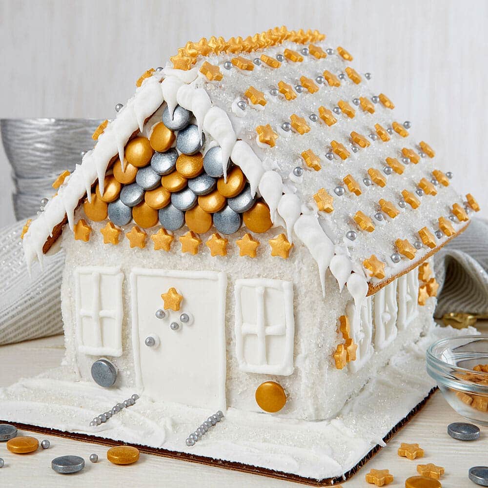 Sparkly white gingerbread house with gold star sprinkles lining the roof. Gold and silver circular sprinkles cover the front of the house above the door with gold stars in a row below.